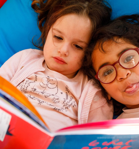 deux fillettes lisent ensemble un livre. L'une d'elles tire la langue vers l'objectif. 