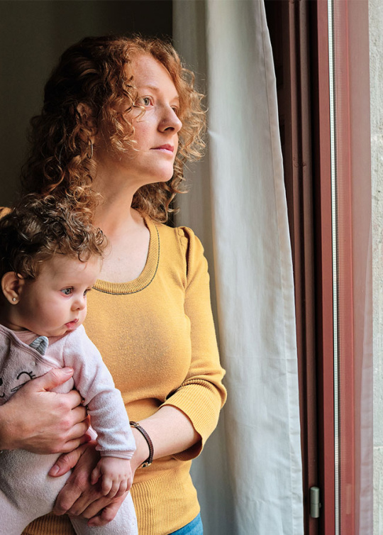 Une maman portant son bébé dans ses bras et regardant par la fenêtre de son appartement