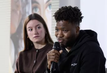 Un jeune homme parle au micro. A sa droite, une jeune fille le regarde. 