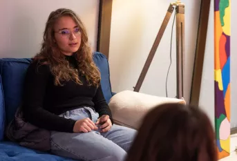Jeune femme assise sur un canapé, de face. On aperçoit au 1e plan le dos de la tête de son interlocutrice. 