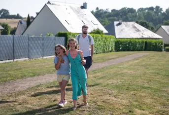 Deux petites filles au 1e plan, 1 adulte un peu plus loin, sont dehors sur un chemin herbeux devant des bâtiments. 