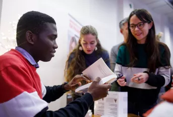 Un jeune homme assis dédicace un livre à une jeune femme debout. Derrière elle, d'autres personnes font la queue.