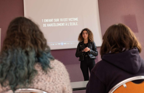 Devant deux jeunes de dos, une jeune femme se tient devant un écran de projection sur lequel est écrit "1 enfant sur 10 est victime de harcèlement à l'école".  