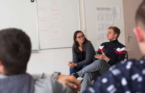 La photo montre Béatrice et un jeune lycéen de face dans une salle de classe. On aperçoit d'autres personnes en bord cadre. 