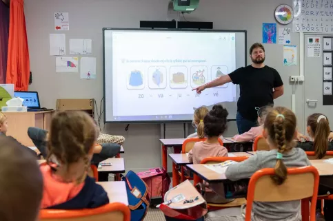 Des élèves de primaire (vus de dos) sont face à leur professeur des écoles. 