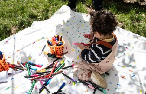 Installée sur une nappe posée sur de l'herbe, un enfant joue avec des crayons de couleur