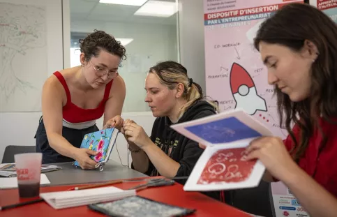 2 jeunes femmes travaillent des reliures assises à une table. Une 3e femme est debout et montre comment faire. 