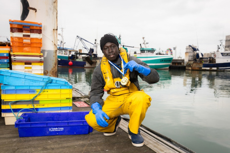 Un jeune homme en tenue de pêcheur sur un quai, une médaille autour du cou