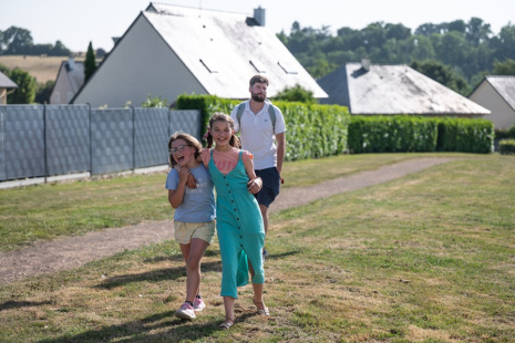 Deux petites filles au 1e plan, 1 adulte un peu plus loin, sont dehors sur un chemin herbeux devant des bâtiments. 